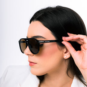 Modelo utilizando óculos de sol feminino Rosa Coral Sassari, armação Preta e lente Marrom– estilo sofisticado e proteção UVA e UVB 400 para os olhos.